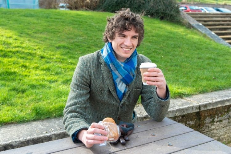 Student sitting on a bench with a doughnut in his right hand and coffee cup in his left hand