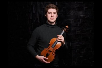 Violinist Michael Foyle stands against a black background holding a violin. He is wearing a black jumper.