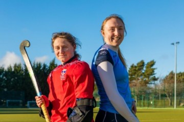 Two hockey players in hockey gear stand back to back on a hockey pitch against a blue sky. Both are holding their sticks, one is in goal-keeper kit.