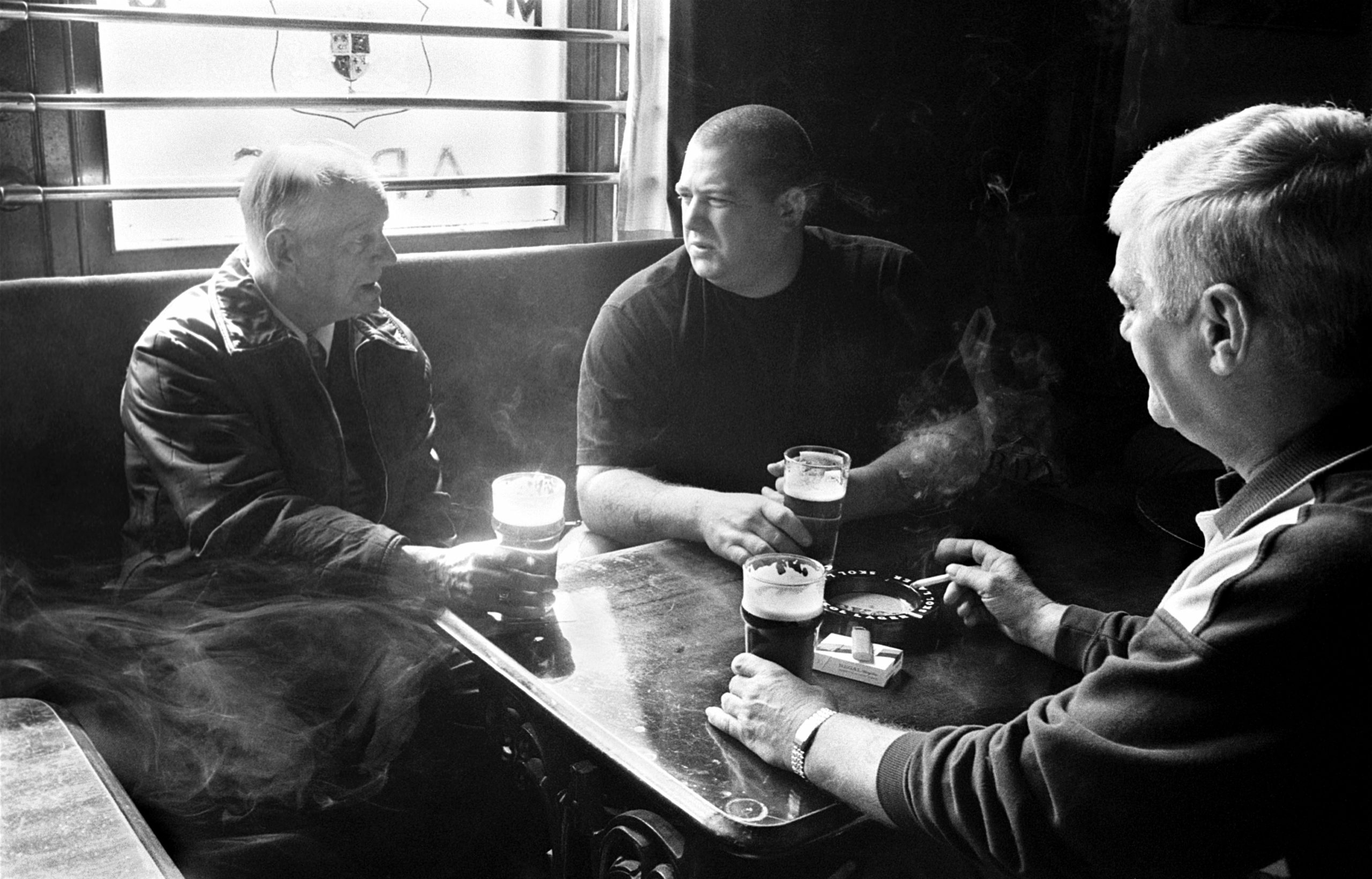 Three Scottish miners sit round a pub table drinking and smoking, light filters in the window. Image is black and white.