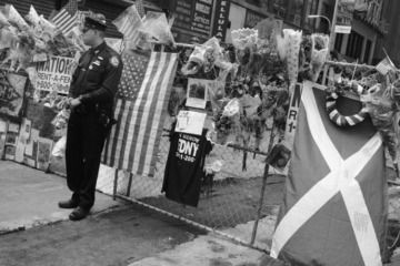 Black and white image of a New York policeman standing in front of a fence covered in flowers and flags following the events of 9/11. A saltire is draped on the fence.