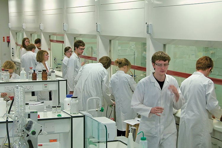 Undergraduate students in white lab coats are working at benches in a laboratory