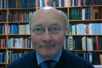 A man - Dr Tim Greenwood - wearing glasses sits in front of a bookshelf full of books