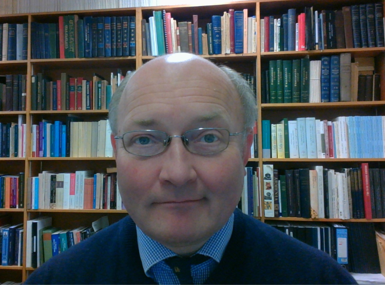 A man - Dr Tim Greenwood - wearing glasses sits in front of a bookshelf full of books