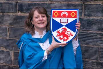 woman in blue academic gown holds up university crest