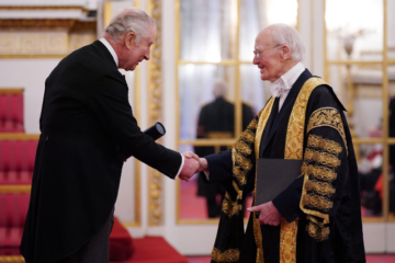 King Charles, holding a scroll in a tube, shakes the hand of Lord Campbell of Pittenweem, who is wearing University robes. The red carpet and mirrored door of Buckingham Palace can be seen in the background