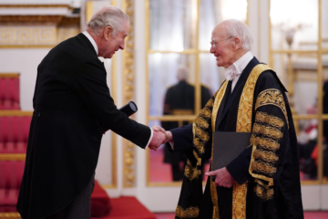 King Charles, holding a scroll in a tube, shakes the hand of Lord Campbell of Pittenweem, who is wearing University robes.