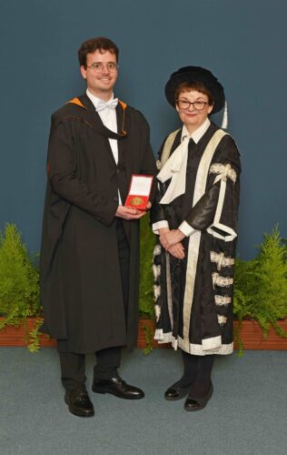A man holding a medal stands next to the Principal in her robes